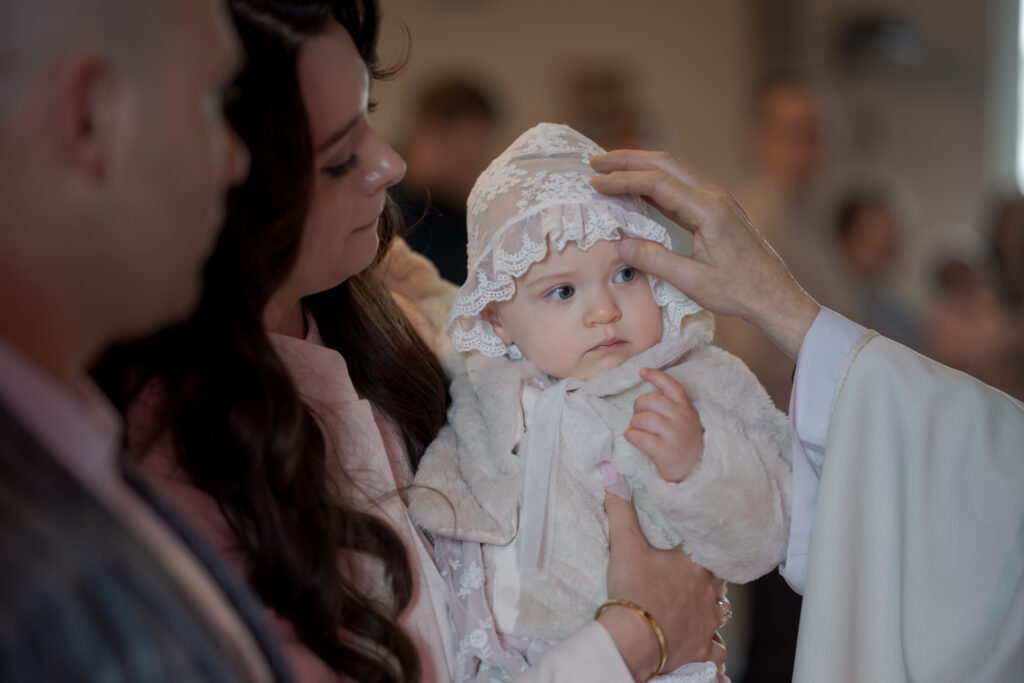 Duchowny błogosławi niemowlę trzymane przez rodziców podczas chrztu